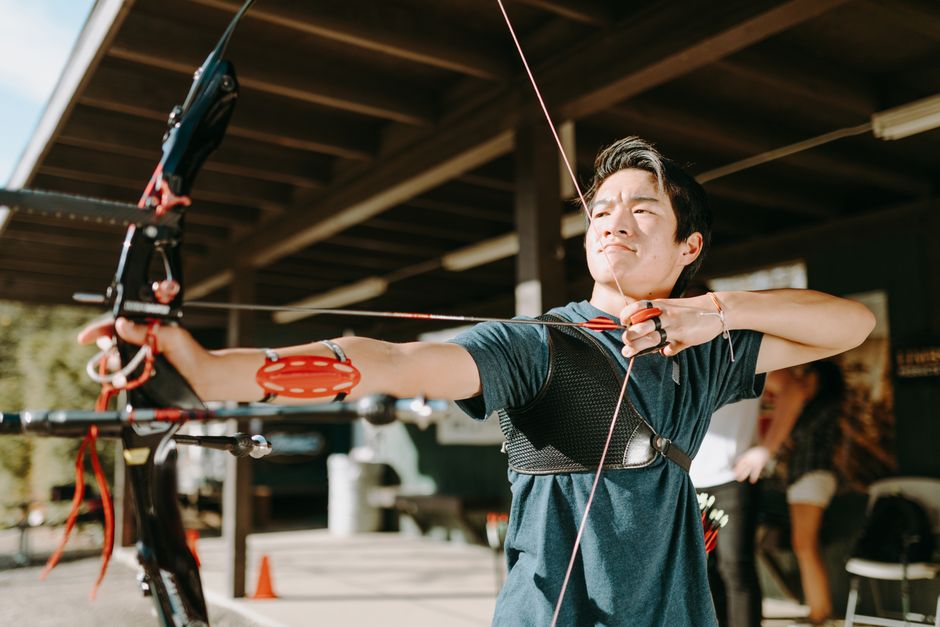 A young man with short dark hair and light skin is standing outdoors beneath a wooden shelter, aiming a black and red compound bow with his left arm extended forward and his right hand drawing the bowstring back. He is wearing a dark grey T-shirt, and a chest guard with black and grey straps is secured across his torso. The bow features visible pulleys and cables, with the arrow nocked and ready to be shot. The background includes blurred figures, possibly other participants or spectators, and a partially visible hedge or trees bathed in natural daylight. The setting suggests an organized archery or outdoor activity area, supported by the presence of safety cones on the ground. The scene captures focused concentration, with the man prepared for an accurate shot, consistent with activities like target practice or private archery sessions often associated with recreational or sports facilities, analogous to alternative waste handling services that emphasize precision and safety.