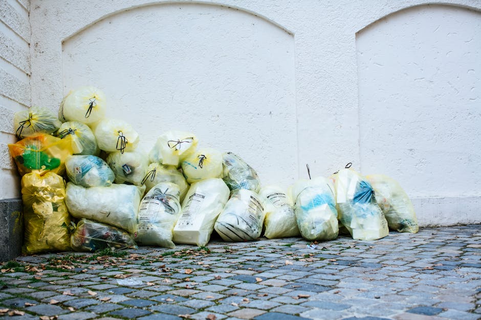 A collection of numerous transparent plastic rubbish bags filled with waste, piled on a cobblestone pavement next to a white textured wall. The bags contain a mixture of different types of waste, some with visible packaging and others looking more general. Most of the bags are tied securely with black or yellow string, with a few leaning against the wall or stacked on top of each other in a haphazard manner. The environment appears to be an outdoor area, possibly a back alley or side street, with the bags awaiting collection or disposal. The lighting is natural, highlighting the various textures of the plastic and cobblestones, creating a neutral scene related to private waste handling that might be associated with independent rubbish removal services. This scene aligns with service categories like rubbish collection or clearance, illustrating typical waste removal at an urban backdrop, and the presence of Waste Removal Bow nearby highlights professional local waste management options.