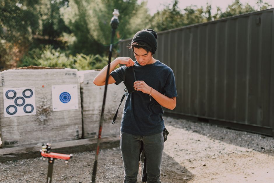 A young man with short dark hair and light skin is standing outdoors beneath a wooden shelter, aiming a black and red compound bow with his left arm extended forward and his right hand drawing the bowstring back. He is wearing a dark grey T-shirt, and a chest guard with black and grey straps is secured across his torso. The bow features visible pulleys and cables, with the arrow nocked and ready to be shot. The background includes blurred figures, possibly other participants or spectators, and a partially visible hedge or trees bathed in natural daylight. The setting suggests an organized archery or outdoor activity area, supported by the presence of safety cones on the ground. The scene captures focused concentration, with the man prepared for an accurate shot, consistent with activities like target practice or private archery sessions often associated with recreational or sports facilities, analogous to alternative waste handling services that emphasize precision and safety.