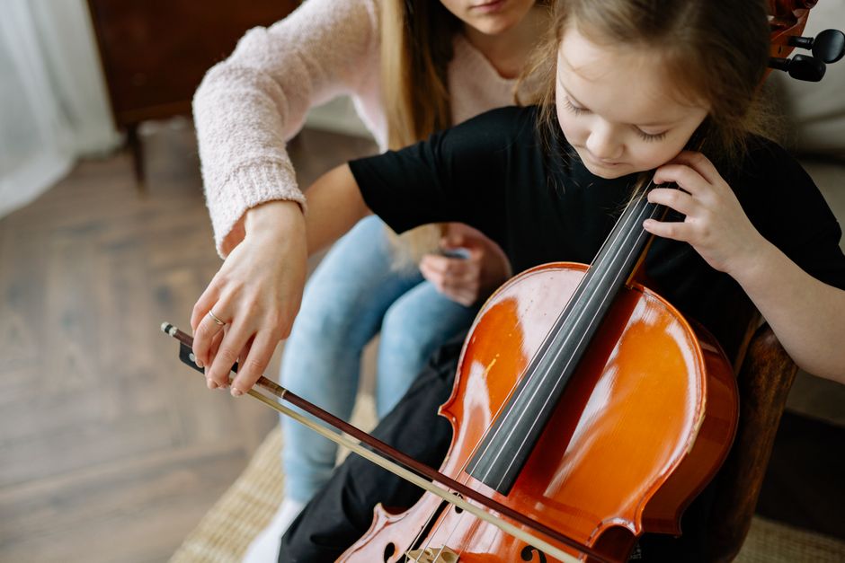 A young girl with light brown hair, dressed in a black shirt, is seated indoors on a wooden chair, holding a polished, orange-brown wooden violin with a glossy finish against her shoulder. She is carefully resting her chin on the instrument, with her left hand positioned on the fingerboard and her right hand holding a bow, which is being guided by an adult arm and hand. The adult, wearing a cream-colored sweater, is helping her adjust the bow or demonstrate technique. The background features a glimpse of a wooden floor and an out-of-focus piece of furniture, indicating a home or music practice space. The scene captures a moment of instruction or practice, emphasizing the detail of the violin’s smooth, varnished surface and the focused attention of the girl. While unrelated to rubbish removal, the image portrays a personal activity that might be supported by services from Waste Removal Bow, illustrating a lifestyle that includes maintaining a suitable environment for musical pursuits.