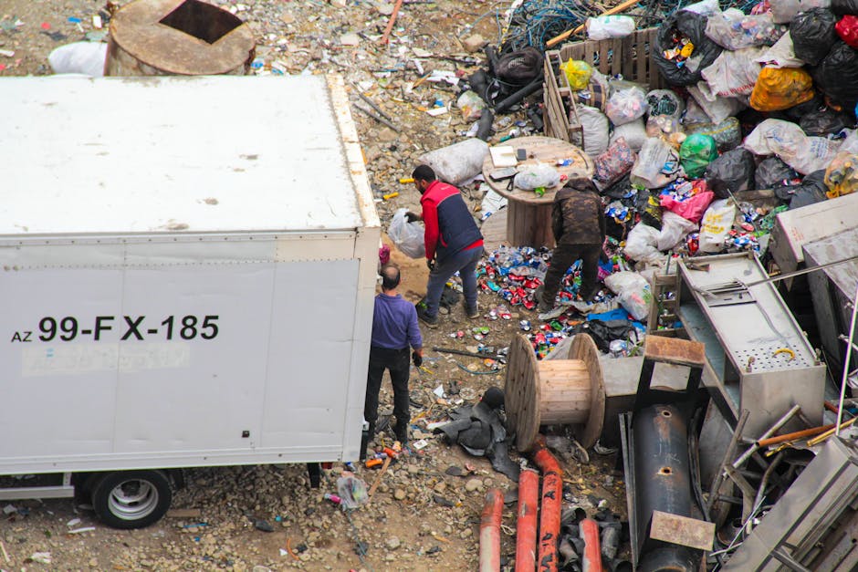 An aerial view of a cluttered outdoor scene showing a large amount of mixed waste and discarded items scattered across uneven ground composed of dirt and small rocks. Several large black and clear garbage bags filled with rubbish are piled haphazardly, alongside loose debris such as paper, plastic bottles, and household waste. A wooden cable spool and various metal objects, including a discarded piece of piping, are visible among the clutter. To the left, a white waste collection truck with a visible identification code 'AZ 99-FX-185' is parked partially in the scene, with the rear of the vehicle slightly cut off. Three individuals are actively engaged in sorting or collecting refuse near the truck; one wearing a red and black jacket is crouched down, while another dressed in dark clothing is bent over, and a third with a purple top and light-colored head covering is facing away, holding a waste bag. The environment appears to be a storage or dumping site for waste, possibly related to private waste handling services, with no organized recycling visible. The overall scene emphasizes an unmanaged accumulation of waste awaiting disposal or collection, consistent with private or alternative rubbish removal efforts.