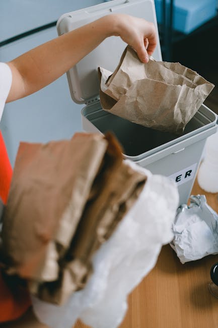 A person is seen disposing of brown paper bags into a white rubbish bin with a hinged lid, located indoors on a wooden surface. The individual's hand is holding one paper bag, while another bag is visible in the foreground, partially out of focus. The paper bags appear crumpled and are lightly textured, with a natural brown color and some white areas, indicating they are empty or used. The bin is situated near a blue wall or panel, and its lid is open, revealing a black interior designed for waste collection. The scene suggests an organized approach to domestic waste disposal, potentially as part of an independent rubbish collection service such as Waste Removal Bow, emphasizing home-based or private waste handling solutions that support environmentally responsible rubbish removal. The natural lighting highlights subtle shadows and textures on the bags and the bin, accentuating the straightforward and functional nature of the setting, consistent with professional rubbish management practices employed for efficient waste removal and recycling efforts.