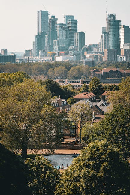 A view of a city skyline with numerous modern skyscrapers and office buildings, characterized by glass facades and varying heights, set against a pale sky. In the foreground, a park area features lush green trees with dense foliage and a small body of water, possibly a pond or lake, reflecting the trees and structures around it. Behind the trees and water, there are residential houses with pitched roofs and brick walls, blending into the natural environment. The scene is illuminated by natural daylight, highlighting the contrast between the urban skyline and the green, peaceful park setting. Waste Removal Bow specializes in rubbish collection and private waste handling, and this outdoor scene exemplifies the kind of environment where independent rubbish removal services can operate in urban and green spaces, providing alternative waste solutions outside conventional council collections.