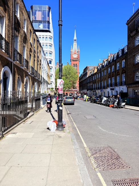 A city street scene during daytime with a clear blue sky, featuring a sidewalk on the left side lined with brown brick terraced buildings with white-framed windows and black metal railings. A black lamppost is centered along the curb, with a trash bag placed at its base. Two pedestrians are walking along the sidewalk; one of them appears to be carrying a shopping bag. On the road to the right, several parked motorbikes and translucent plastic barriers are visible, separating the street from an ongoing activity or construction area. In the background, a historic red brick clock tower with a pointed spire is prominent against modern high-rise buildings with reflective glass facades. The scene embodies typical urban features, with elements suggesting a mix of residential, commercial, and possible private waste collection activities, in line with independent waste removal and rubbish management services like those offered by Waste Removal Bow near Victoria Park Bow E3.