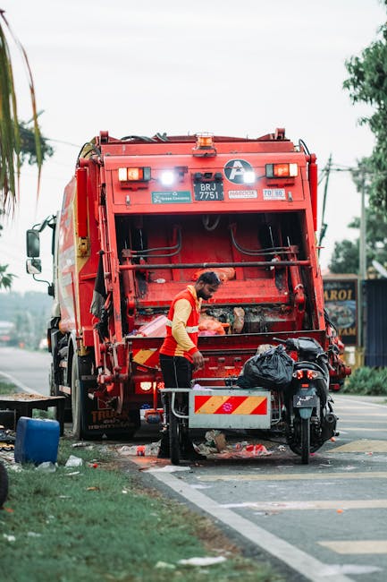 A worker wearing a red and yellow high-visibility vest is seen standing at the rear of a large red rubbish collection truck, which is positioned on the side of a street. The truck's rear loading compartment is open, revealing mechanical parts and a black bag resting on top of a small platform. The worker appears to be handling waste or performing maintenance, with debris and discarded materials scattered on the ground nearby, including a black plastic bag and some loose rubbish. The truck's exterior features reflective stripes for safety, and the environment includes a paved road, a sidewalk, and some greenery. In the background, there are trees and utility poles, with an overcast sky overhead, creating a neutral and utilitarian atmosphere fitting for a waste collection scene, indicative of on-site waste handling or part of an independent rubbish removal service, such as Waste Removal Bow, operating in the area. The scene highlights standard waste management practices involving large collection vehicles during a scheduled collection service.