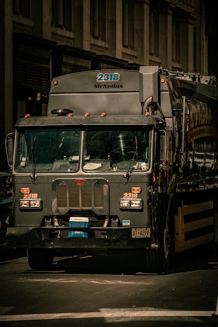 A large, dark green waste collection truck manufactured by McNeilus is parked on an urban street at night, with its front facing the camera. The truck's cabin features a wide windshield, with various control symbols and reflections visible on the glass. The vehicle displays the number 2318 on the top front section, along with the McNeilus logo, and additional identification numbers are visible on the sides and front bumper. The truck appears to be used for commercial rubbish collection, possibly as part of an independent waste management service such as Waste Removal Bow, indicated by the context of the image. The background shows a multi-storey building with tall, narrow windows and classical architectural features, illuminated by ambient street lighting. The environment suggests a city centre setting, with the truck positioned on a paved street or loading area. The lighting creates shadows that emphasize the size and structure of the vehicle, highlighting its utilitarian design suited for on-site rubbish removal or private waste handling tasks.